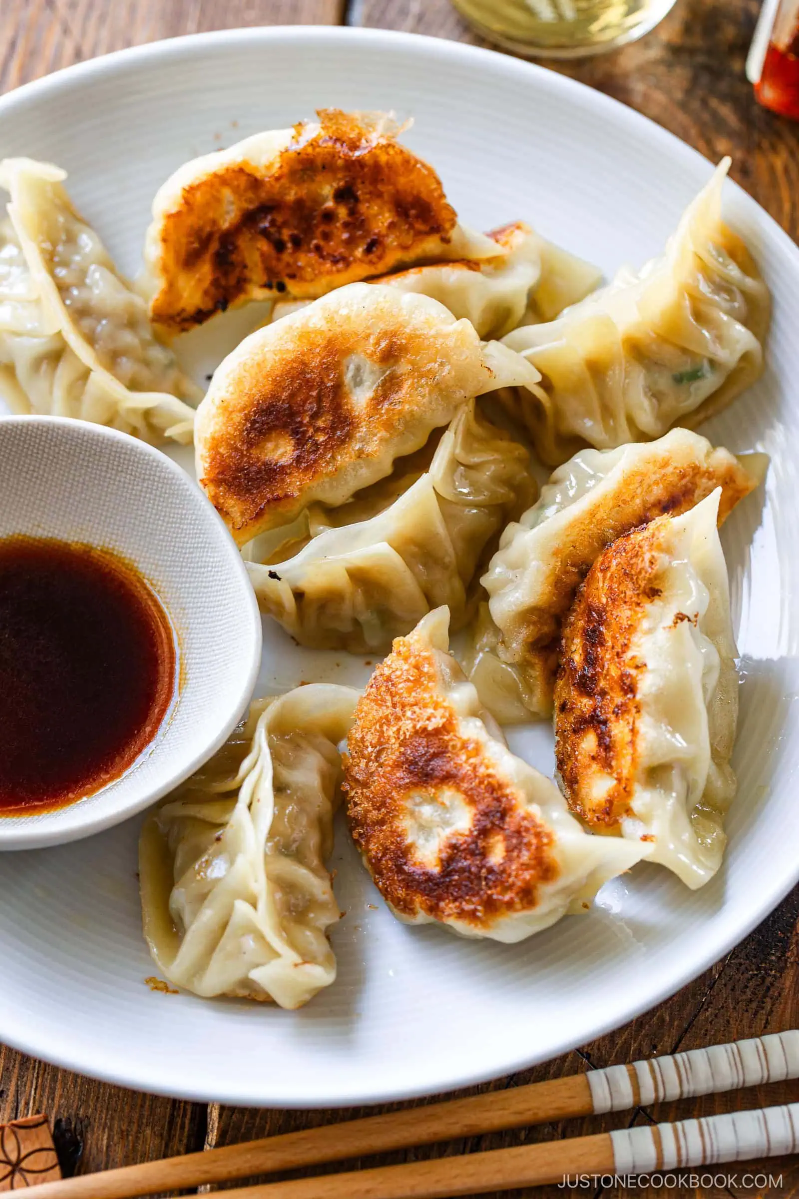 A white plate filled with pan-fried dumplings from a classic gyoza recipe, some with crispy golden bottoms, sits next to a small bowl of dipping sauce and a pair of wooden chopsticks.