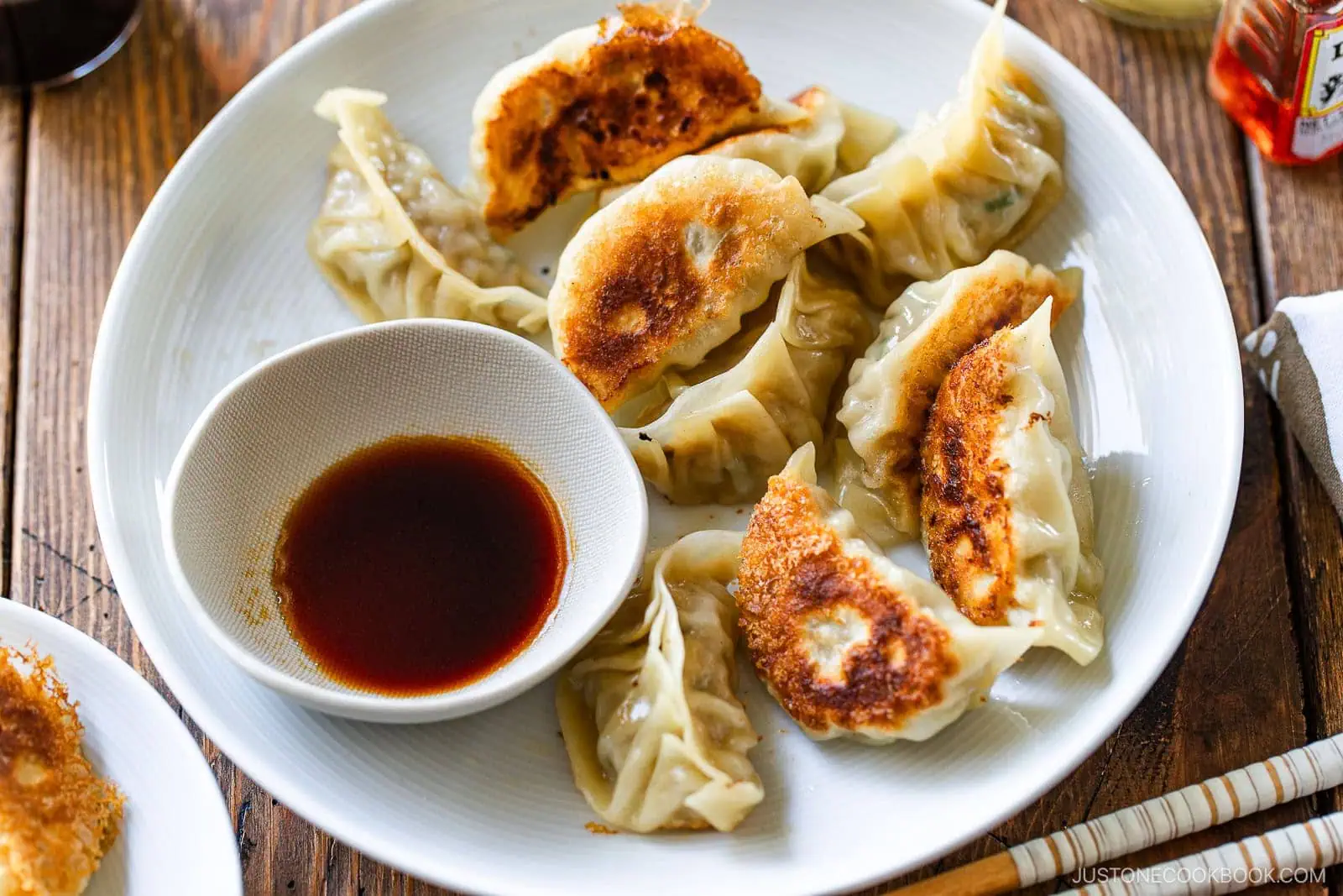 A white plate with golden-brown pan-fried dumplings, made from a classic gyoza recipe, arranged around a small bowl of soy dipping sauce, set on a wooden table.