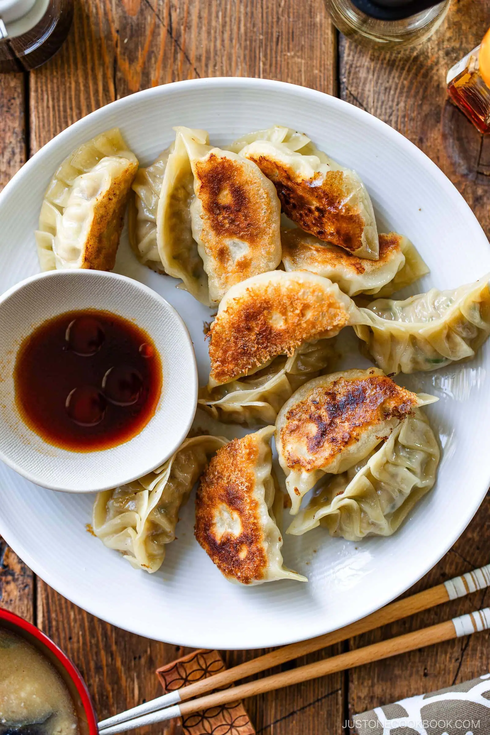 A white plate filled with crispy, golden brown dumplings made from a classic gyoza recipe is served with a small bowl of soy sauce. Chopsticks rest beside the plate on a wooden table.