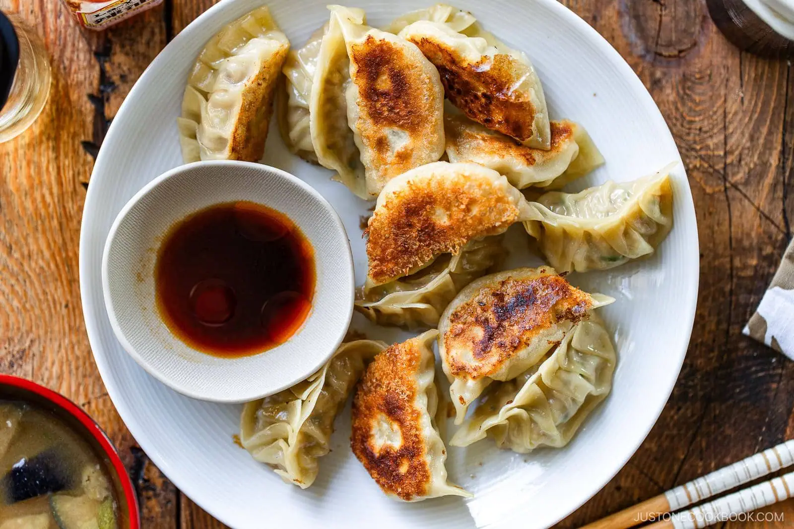 A white plate with pan-fried dumplings from a classic Gyoza Recipe is arranged around a small bowl of soy sauce on a wooden table. Some have crispy golden-brown bottoms, and a bowl of soup is partially visible in the corner.