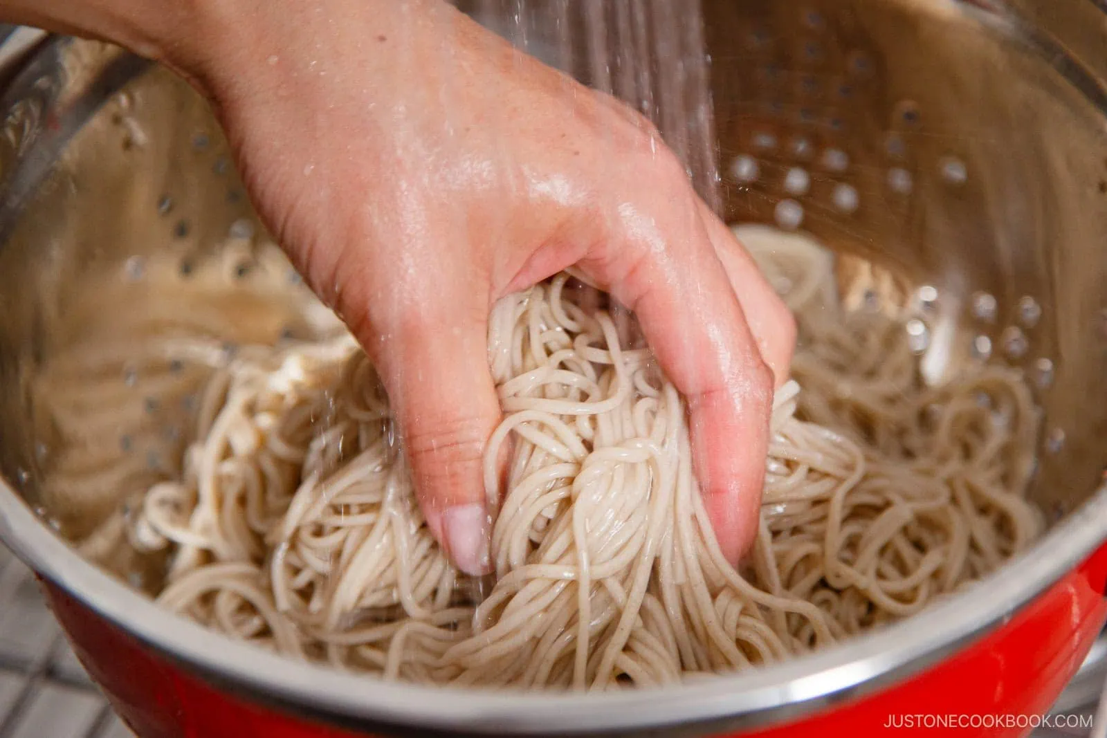 A hand rinsing cooked zaru soba noodles under running water in a metal colander.