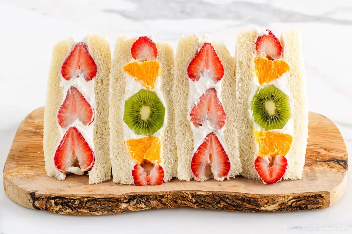 Four Japanese Fruit Sandwiches with cream, filled with slices of strawberries, oranges, and kiwis, are neatly arranged on a wooden board against a light background.