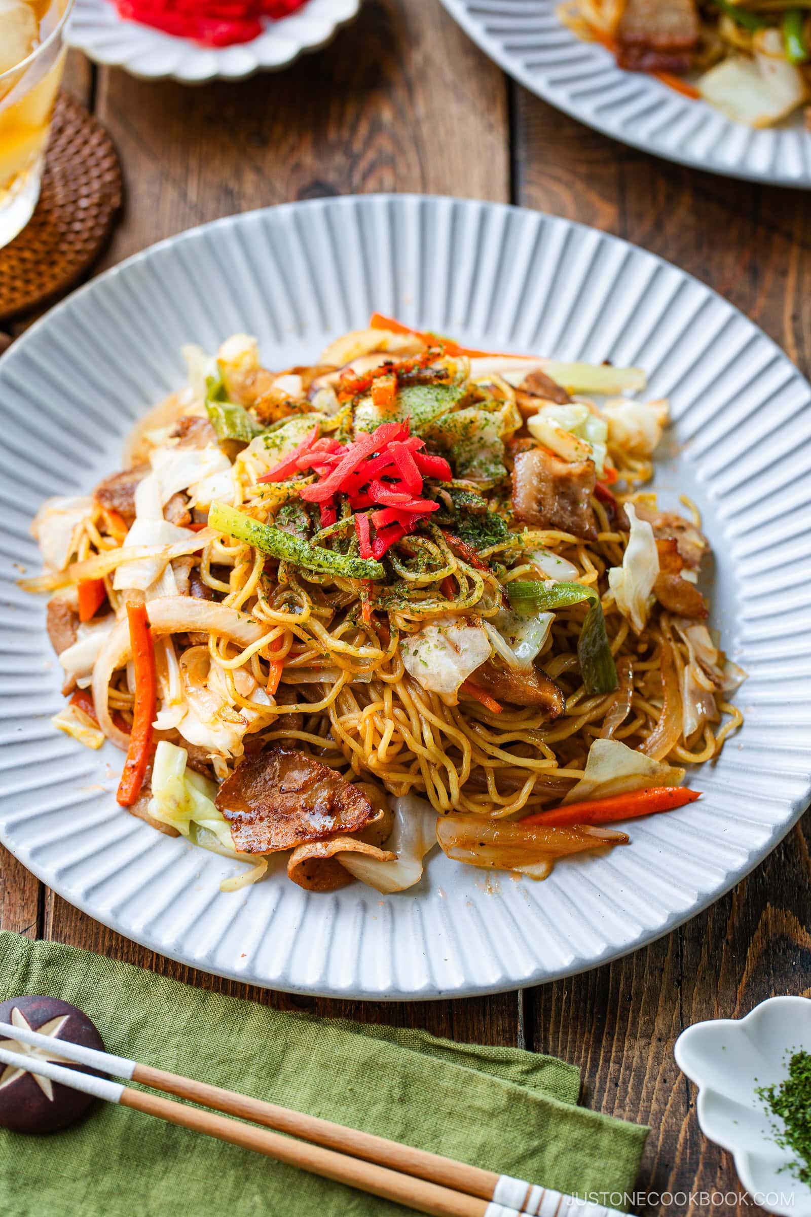 A plate of yakisoba (Japanese stir-fried noodles) with sliced pork, cabbage, and carrots, topped with green seaweed powder and red pickled ginger, served on a white dish with chopsticks and a green napkin on a wooden table.