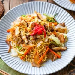 A plate of yakisoba (Japanese stir-fried noodles) topped with sliced pork, vegetables, and pickled ginger, served on a white scalloped plate with chopsticks and a drink on a rustic wooden table.