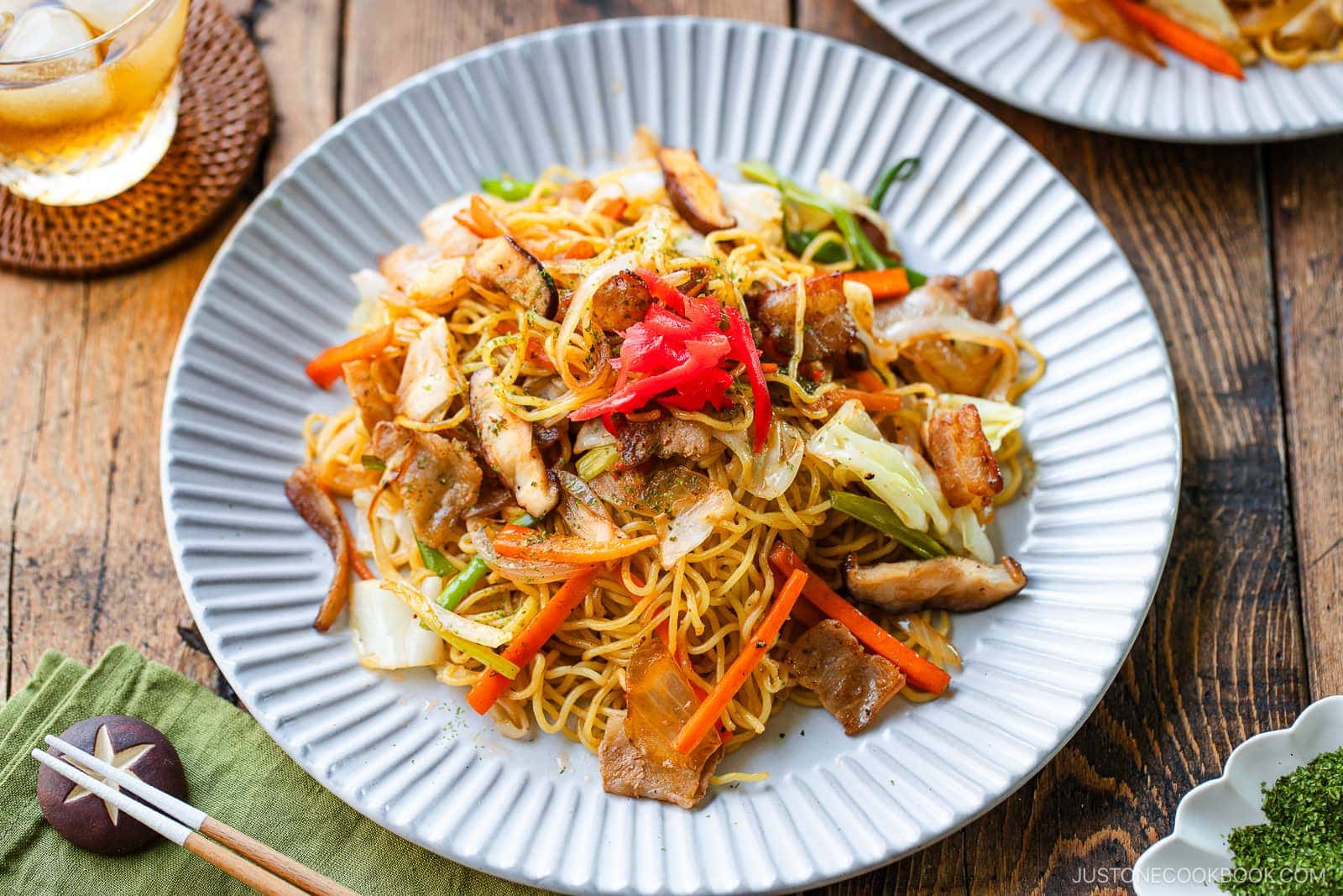A plate of yakisoba (Japanese stir-fried noodles) topped with sliced pork, vegetables, and pickled ginger, served on a white scalloped plate with chopsticks and a drink on a rustic wooden table.