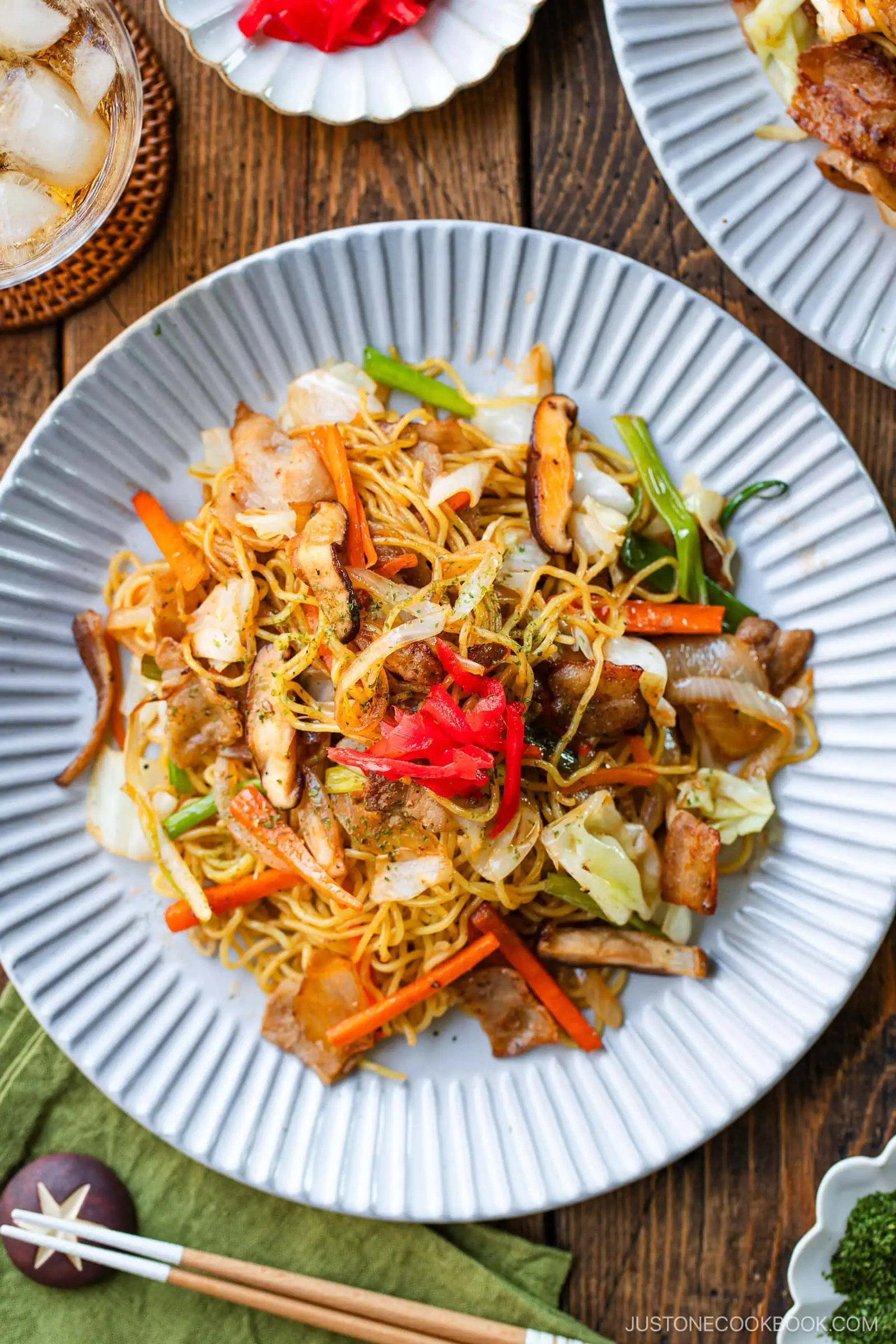 A plate of yakisoba, Japanese stir-fried noodles, topped with vegetables, sliced meat, and red pickled ginger is served on a white plate with a drink and additional dishes in the background.