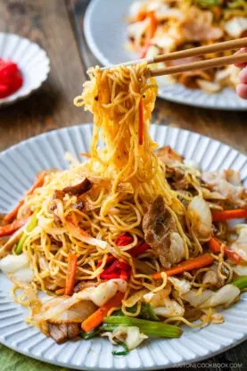 A close-up of chopsticks lifting a portion of yakisoba (Japanese stir-fried noodles) with sliced pork, cabbage, carrots, and green onions on a white plate. Another plate of noodles is visible in the background.