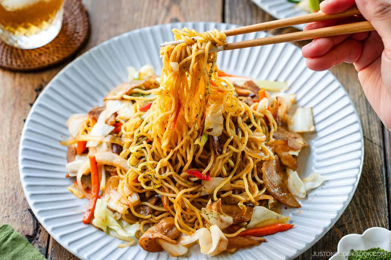A hand holding chopsticks lifts a portion of yakisoba (Japanese stir-fried noodles) with vegetables and meat from a white plate on a wooden table.