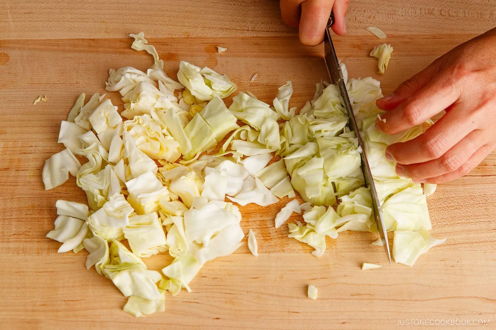 A person&rsquo;s hand is chopping fresh green cabbage on a wooden cutting board with a knife, preparing it for yakisoba. The cabbage is cut into small pieces, and some leaves are scattered around.