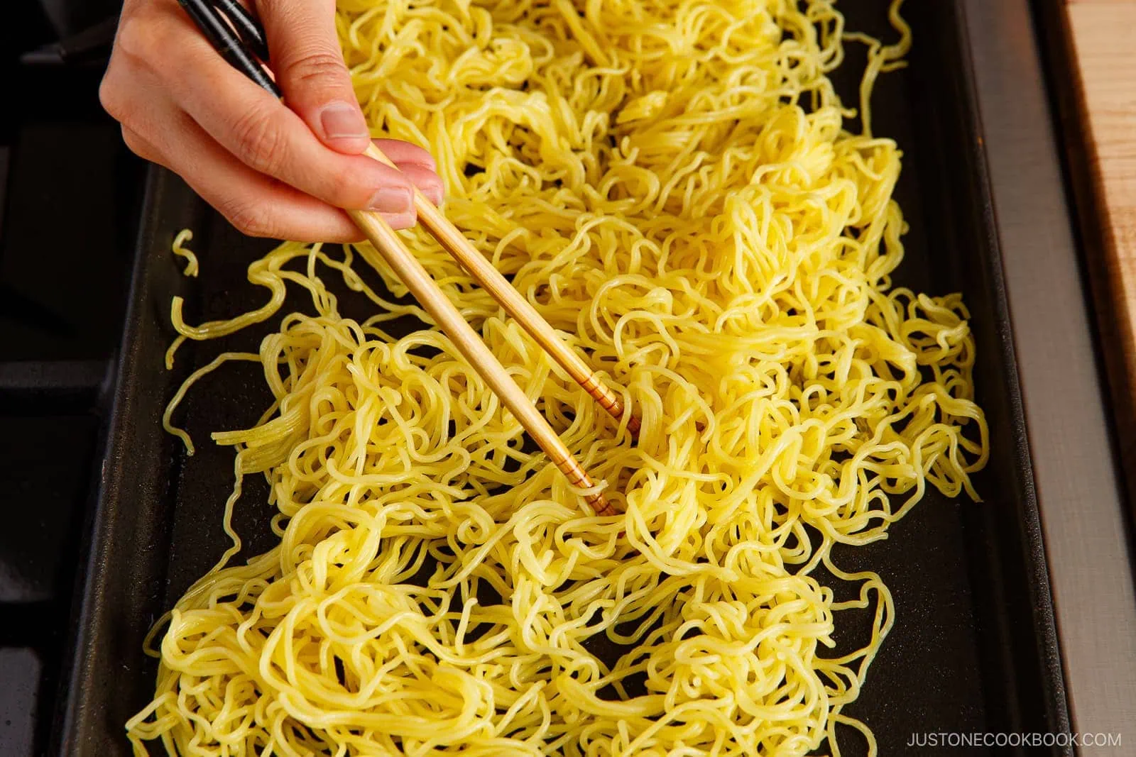 A hand using chopsticks to spread and stir yellow yakisoba (Japanese stir-fried noodles) as they cook on a flat griddle or pan.