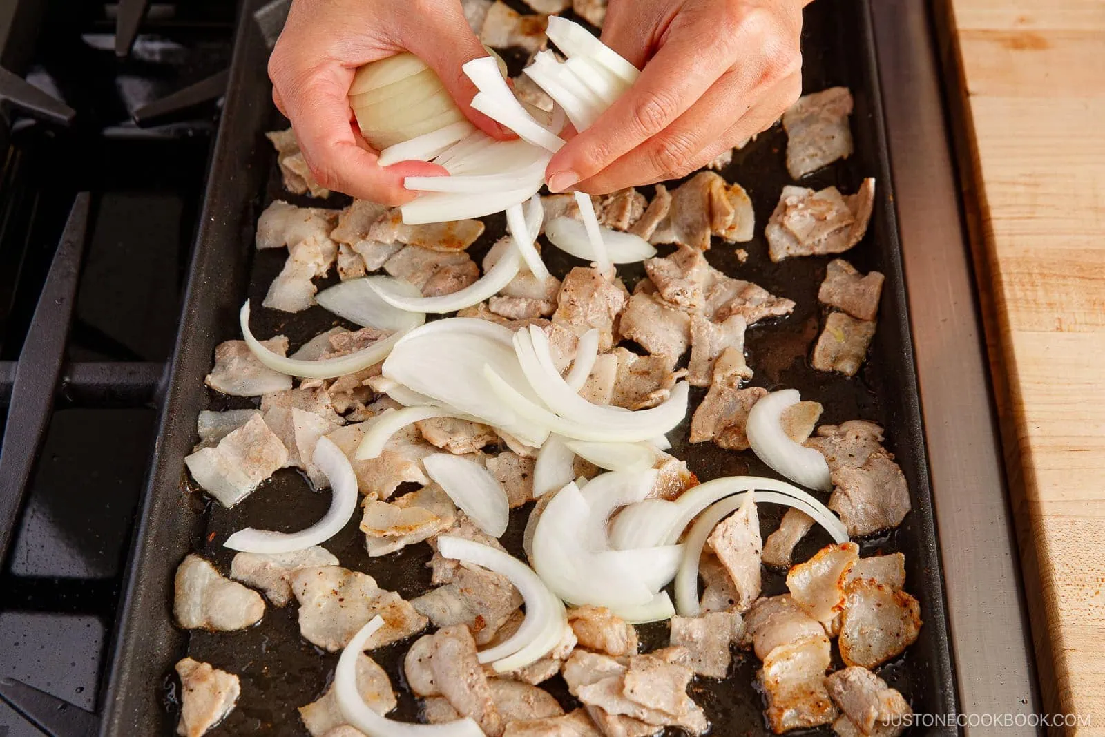 Hands adding sliced onions to a baking sheet filled with cooked pieces of pork belly on a stovetop, perfect for a savory yakisoba (Japanese stir-fried noodles) dish.