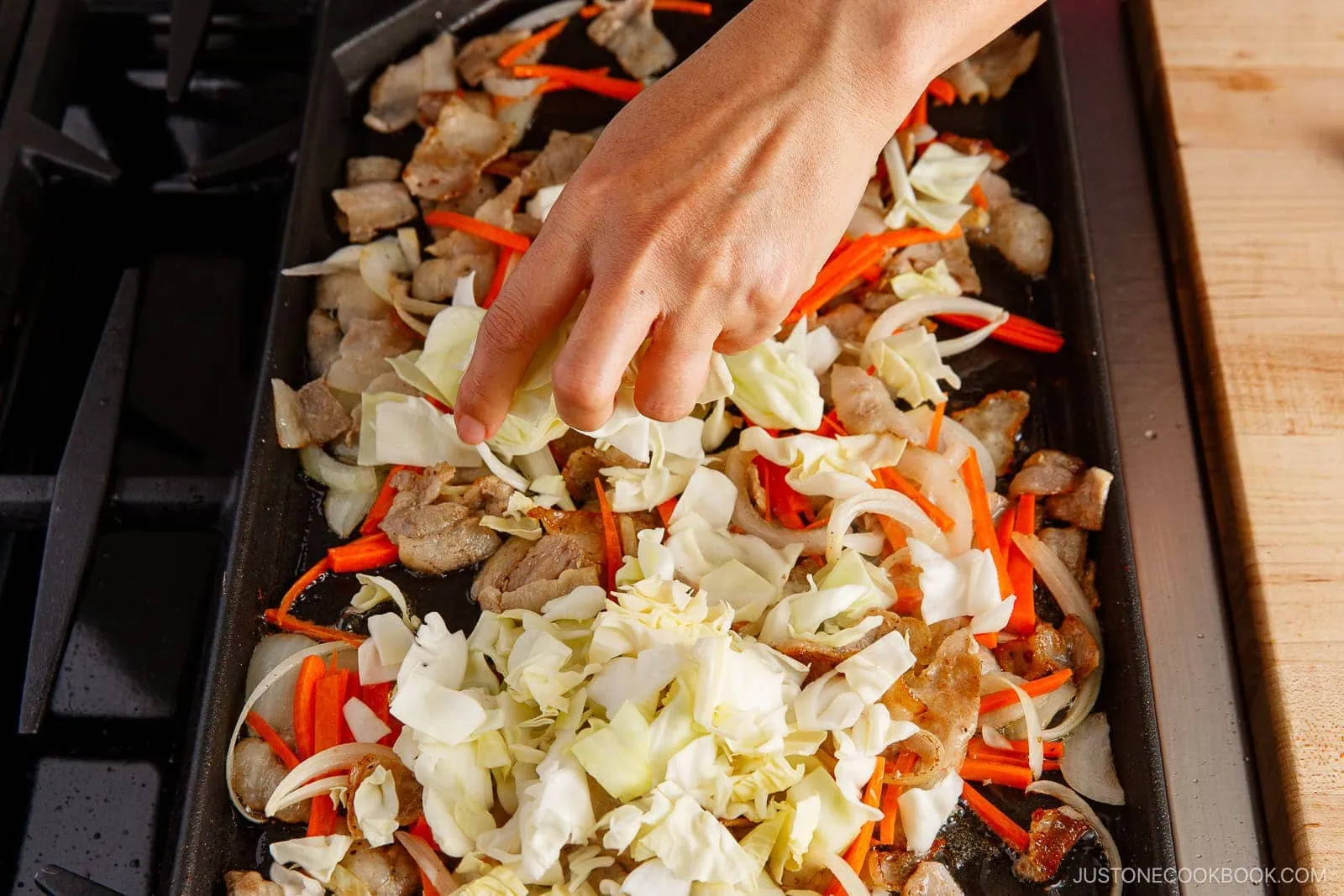 A hand sprinkles chopped cabbage onto a grill pan filled with sliced pork, onions, carrots, and yakisoba, preparing a flavorful Japanese stir-fry on the stovetop.
