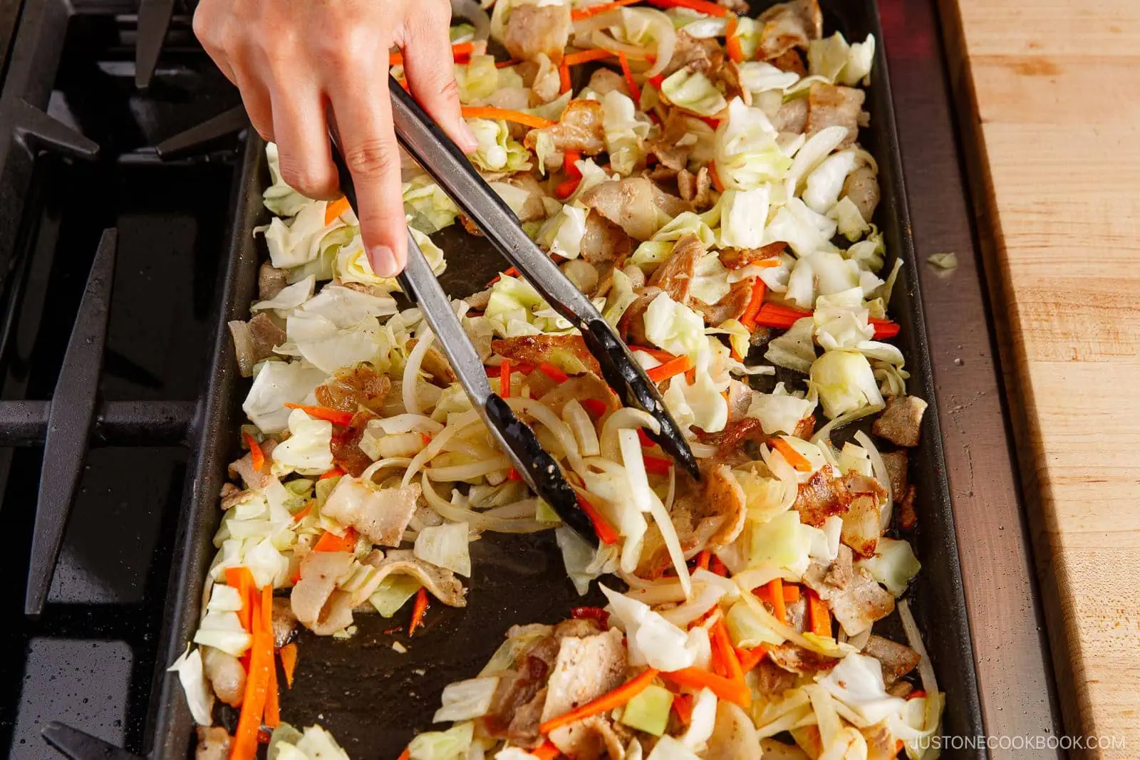 A hand uses tongs to stir-fry chopped cabbage, carrots, onions, meat slices, and yakisoba on a large stovetop griddle. The colorful ingredients and Japanese stir-fried noodles are being cooked evenly across the surface.