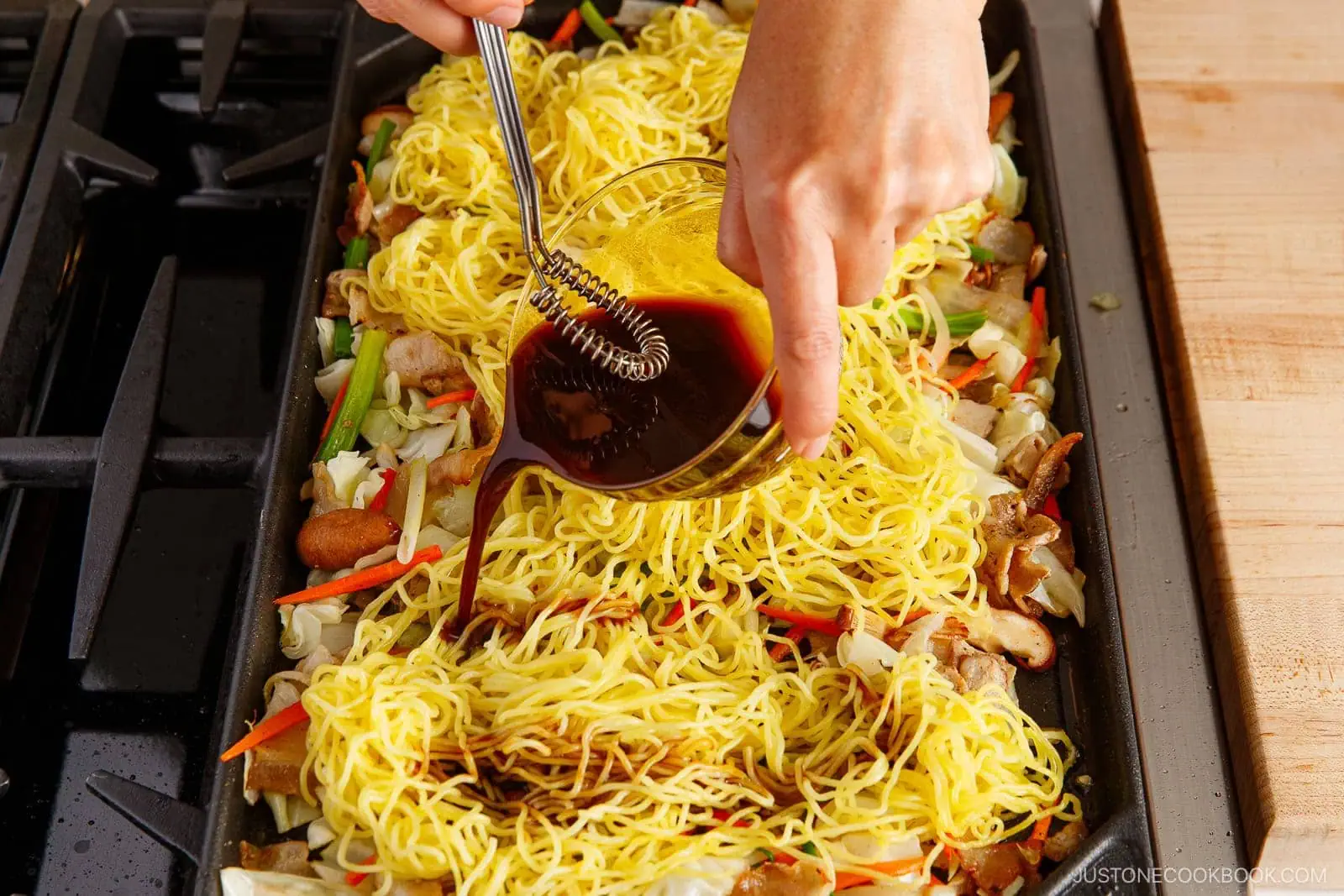 A person pours dark sauce onto yakisoba, a Japanese stir-fried noodle dish with yellow noodles and mixed vegetables cooking on a stovetop griddle.