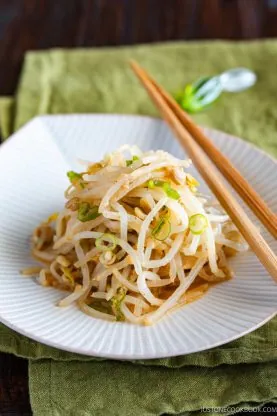A plate of saut&eacute;ed bean sprouts, prepared as a flavorful Bean Sprout Salad and garnished with chopped green onions, is served with wooden chopsticks on a white plate, set atop a green cloth.
